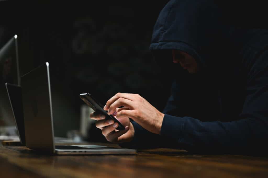 Person using a smartphone beside a laptop in a dark setting, representing Shadow AI and cybersecurity risks for RIAs.